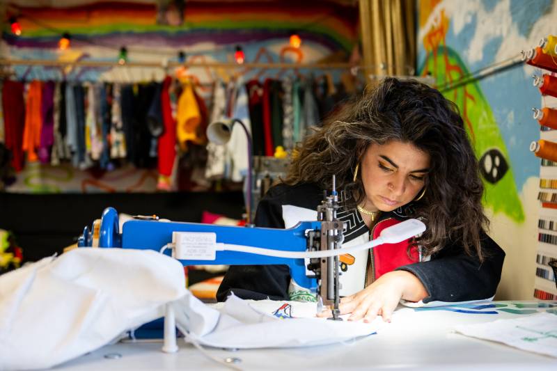 A woman with long wavy hair looks downward as she stitches white fabric with an industrial sewing machine. Colorful garments on a rack hang in the background.