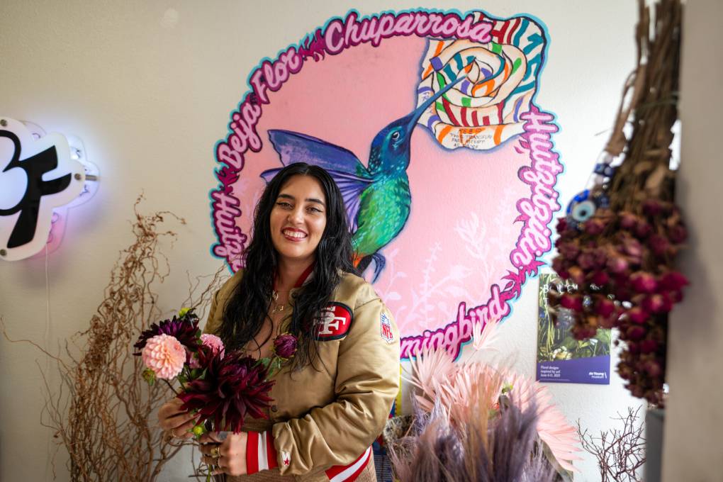 A woman with long hair wears a niners jacket and holds a bouquet of dahlias in front of a hummingbird painted on the wall.