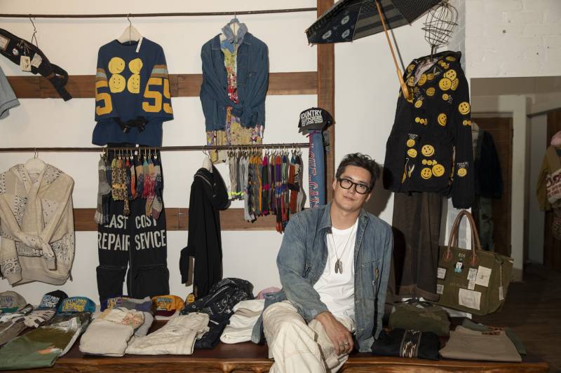 A 32-year-old Asian American man in glasses sits in front of a display of high-end Japanese denim.