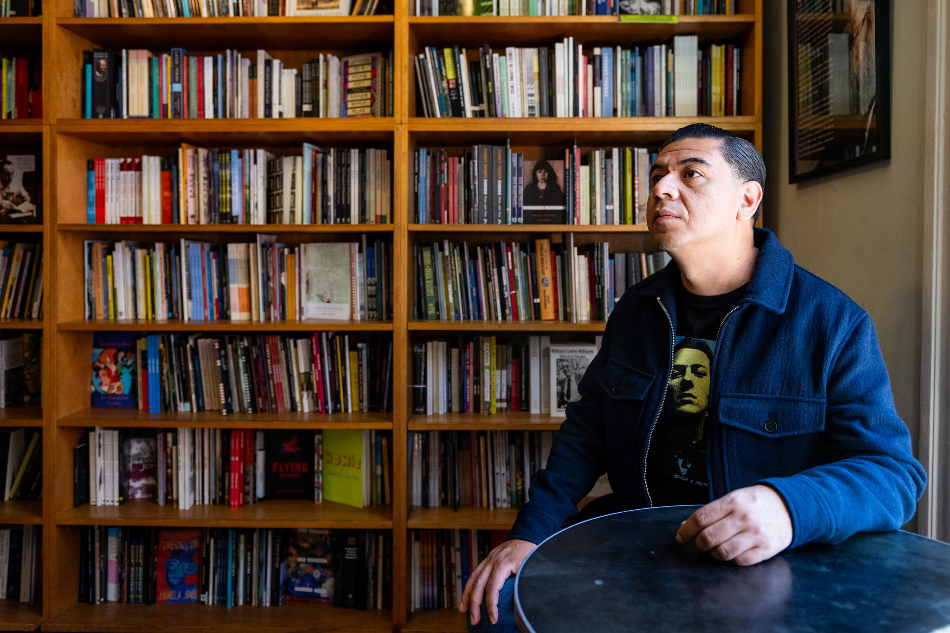 A man in a blue jacket sits inside a bookstore with a full bookshelf behind him.