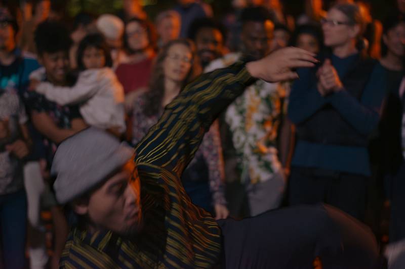 A male dancer does a dance move as a crowd of people watches in the background.