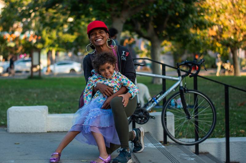 A Black woman wearing a red baseball cap embraces her child in the park while both smile at the camera.
