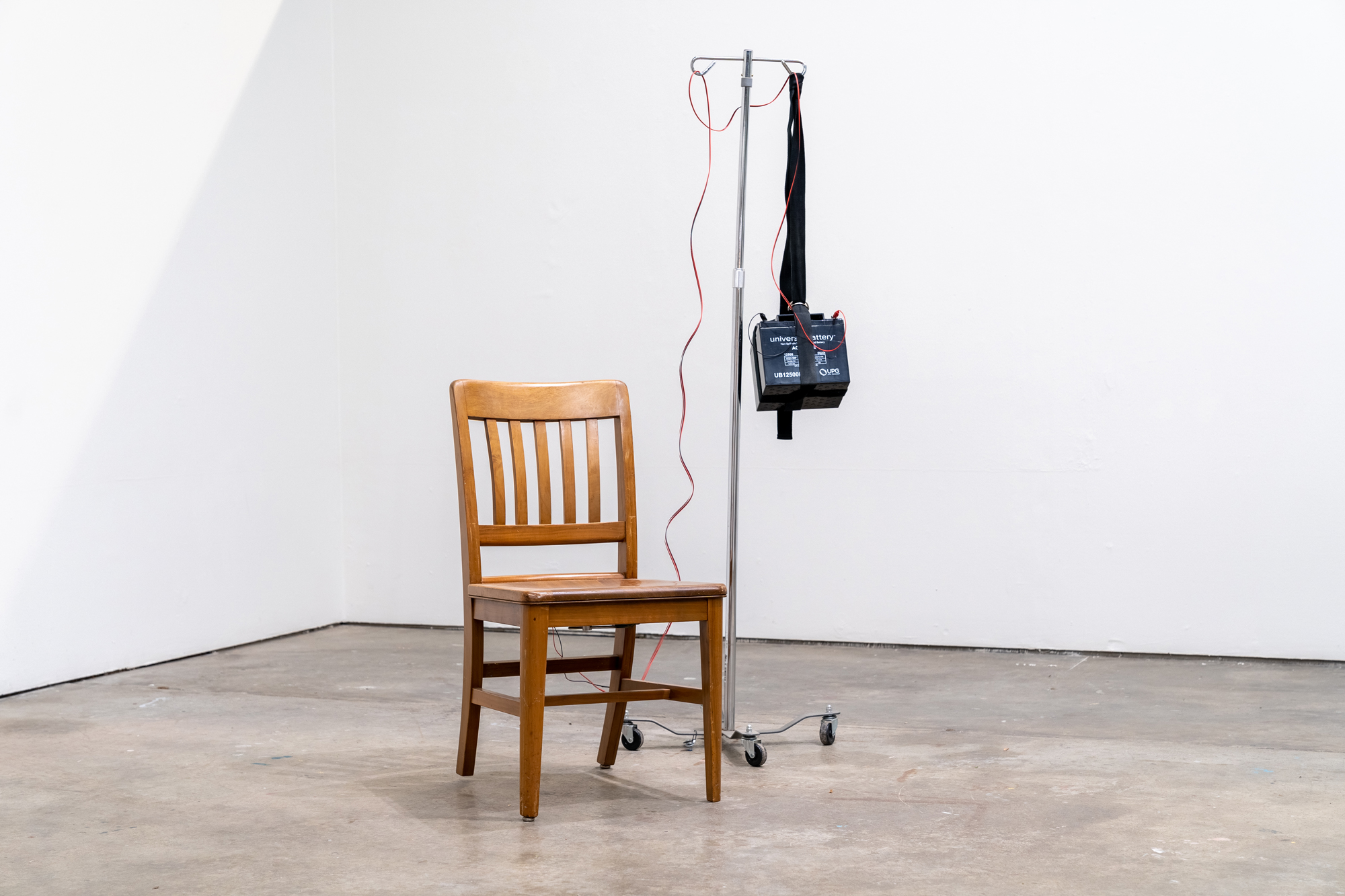 A wooden chair with an IV rack next to it, wire and a car battery hanging from its metal arms