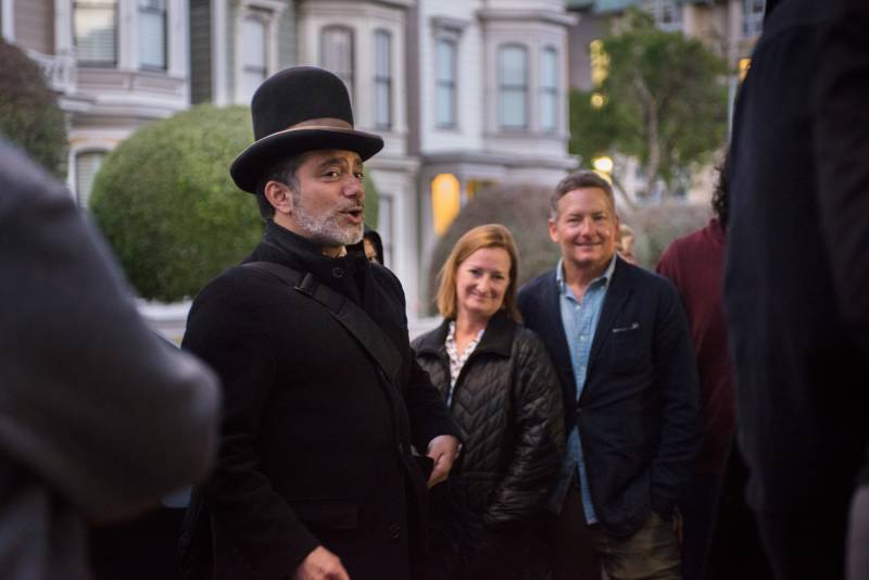 A mixed race man with a short white beard talks to a gathering of people in front of Victorian houses in San Francisco. He is wearing period clothing, including a tall bowler hat and black overcoat.