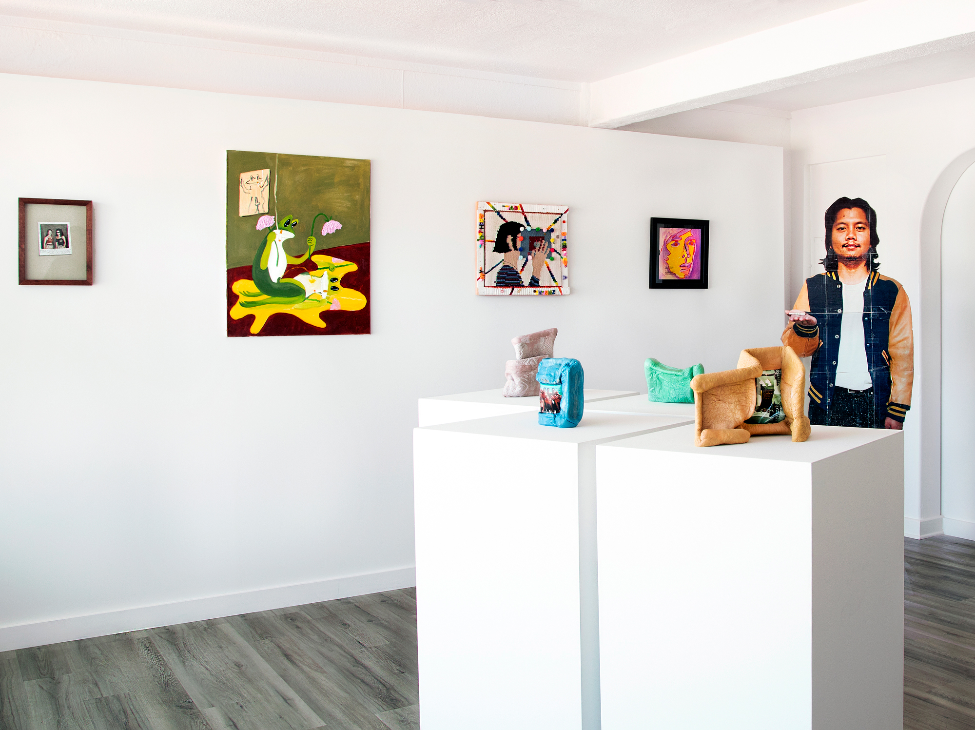 A white gallery space with works on the wall, a life-sized standee of a young man and colorful sculptural works on white pedestals. 