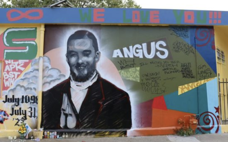 A large scale black and white painting of a young man, on a brick wall. The word Angus is painted in white next to the portrait. On the floor near the mural is a collection of candles.