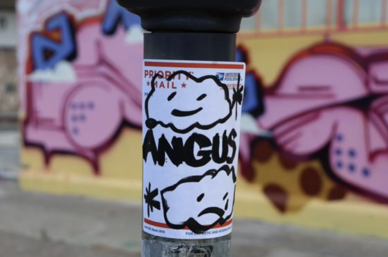 A priority mail address sticker decorated with two clouds - one with a happy face, one shedding a tear - stuck to a post in front of a graffiti'd wall. The clouds have the word ANGUS scrawled between them.