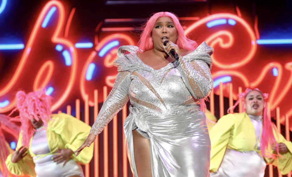 A plus-size Black woman in a pink wig performs on stage in a silver dress with a high slit on one leg. A large neon sign is displayed behind her. Two dancers perform behind her.