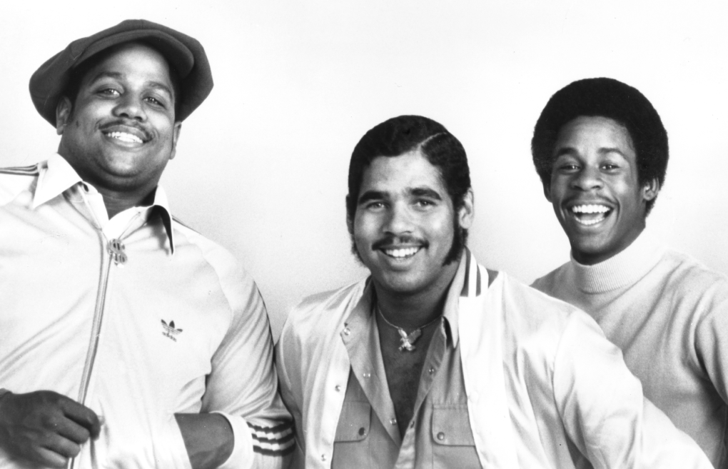 Three Black men stand together smiling. The first wears an Adidas jacket and a peasant hat, the second wears a sports jacket, open shirt and gold necklace, the third is wearing a sweater.