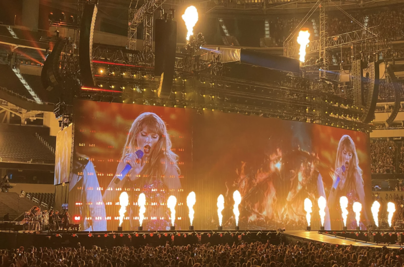 A stadium stage, lit up with columns of flames, shows Taylor Swift performing on large screens.