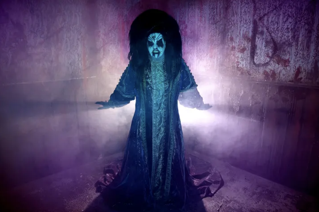 A drag queen wearing white facepaint and elaborate robes stands backlit in a purple foggy room
