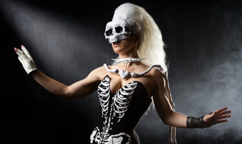 A slender woman in a corset decorated like a human skeleton wears a skull mask and white-blond wig. She is gesturing theatrically.