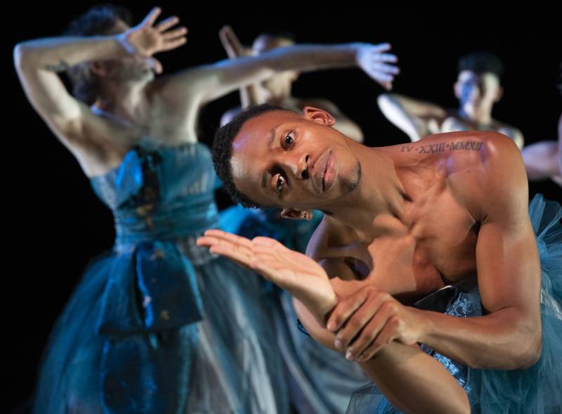 a young Black man dancer poses while facing the camera as other dancers in blue dresses move behind him