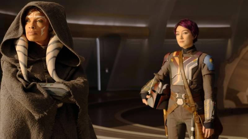 A mixed race woman wearing a hooded cloak and white face paint stands, arms folded, looking off into the distance. A few feet away stands a petite Asian woman wearing a futuristic uniform and holding a helmet. She has short, cropped hair.