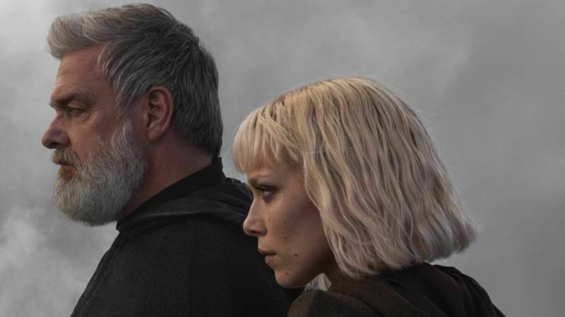 A white man with grey hair and beard stands in profile. Next to him is a petite white woman with white-blonde hair worn in a bob. She has sharp cheekbones.