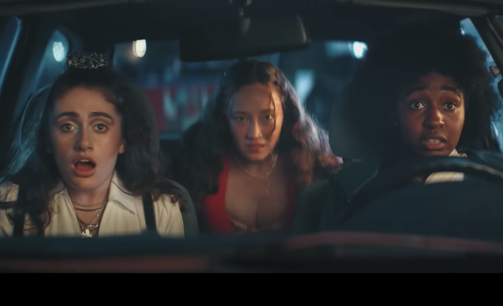 Three girls sit in a car, looking stunning. In the front, a white teen in the passenger seat, a Black teen in the driver's seat and an Asian girl sitting in the back in the center. It is nighttime.