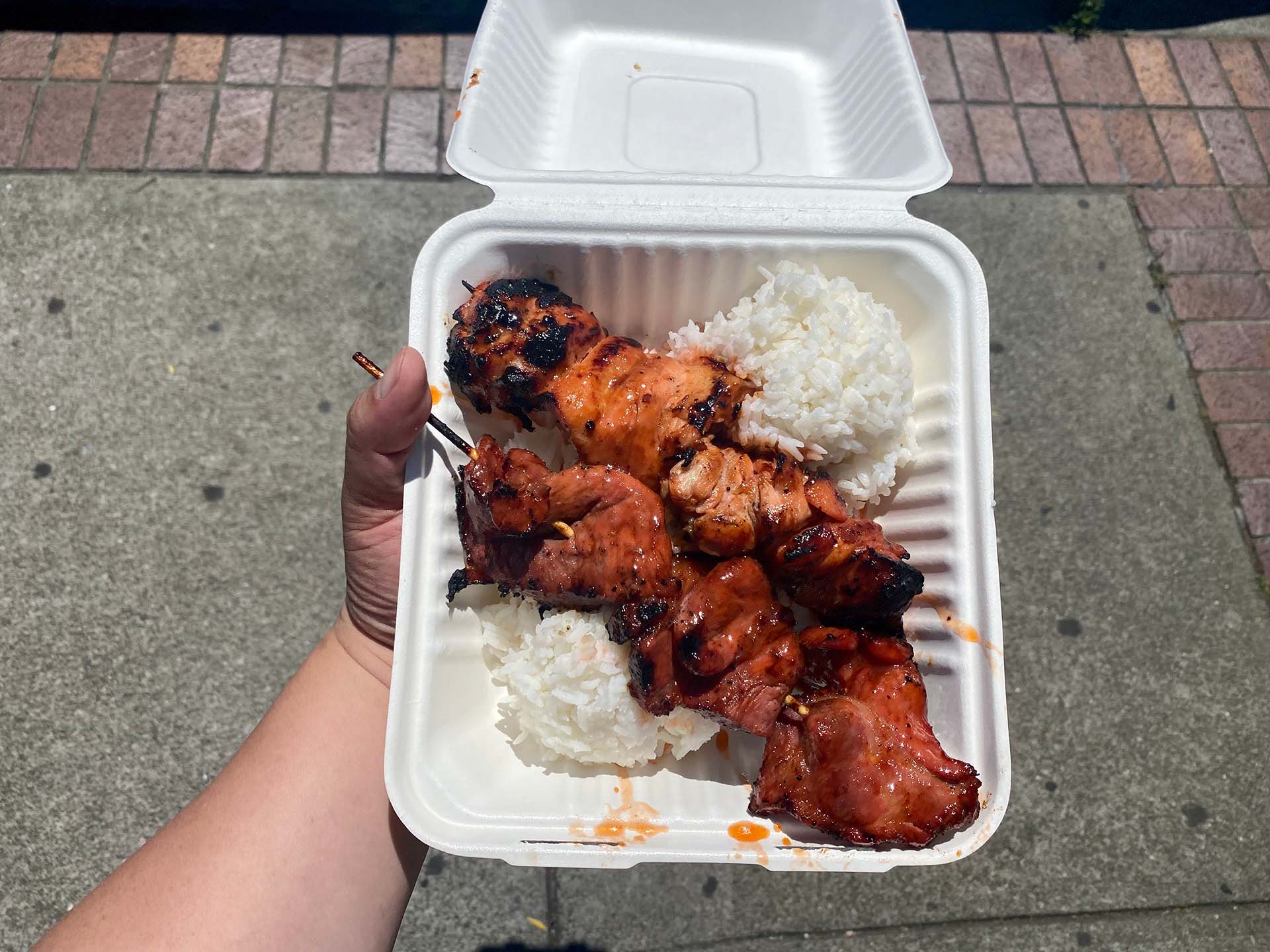 Hand holding a white plastic takeout container of saucy meat skewers arranged over a bed of white rice.