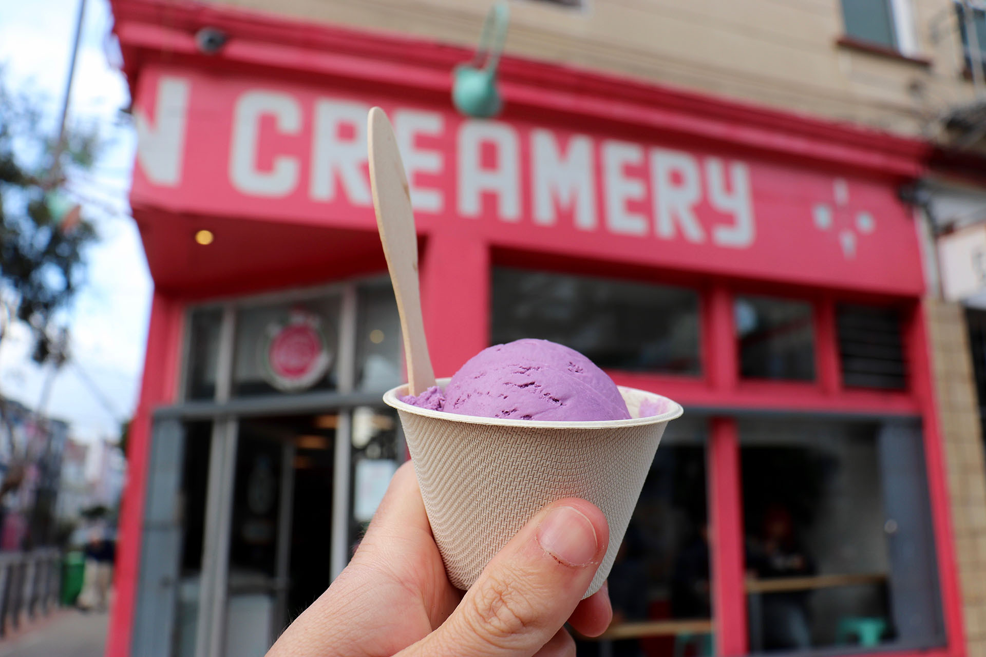 a scoop of purple ice cream is held up in front of a pink storefront in San Francisco