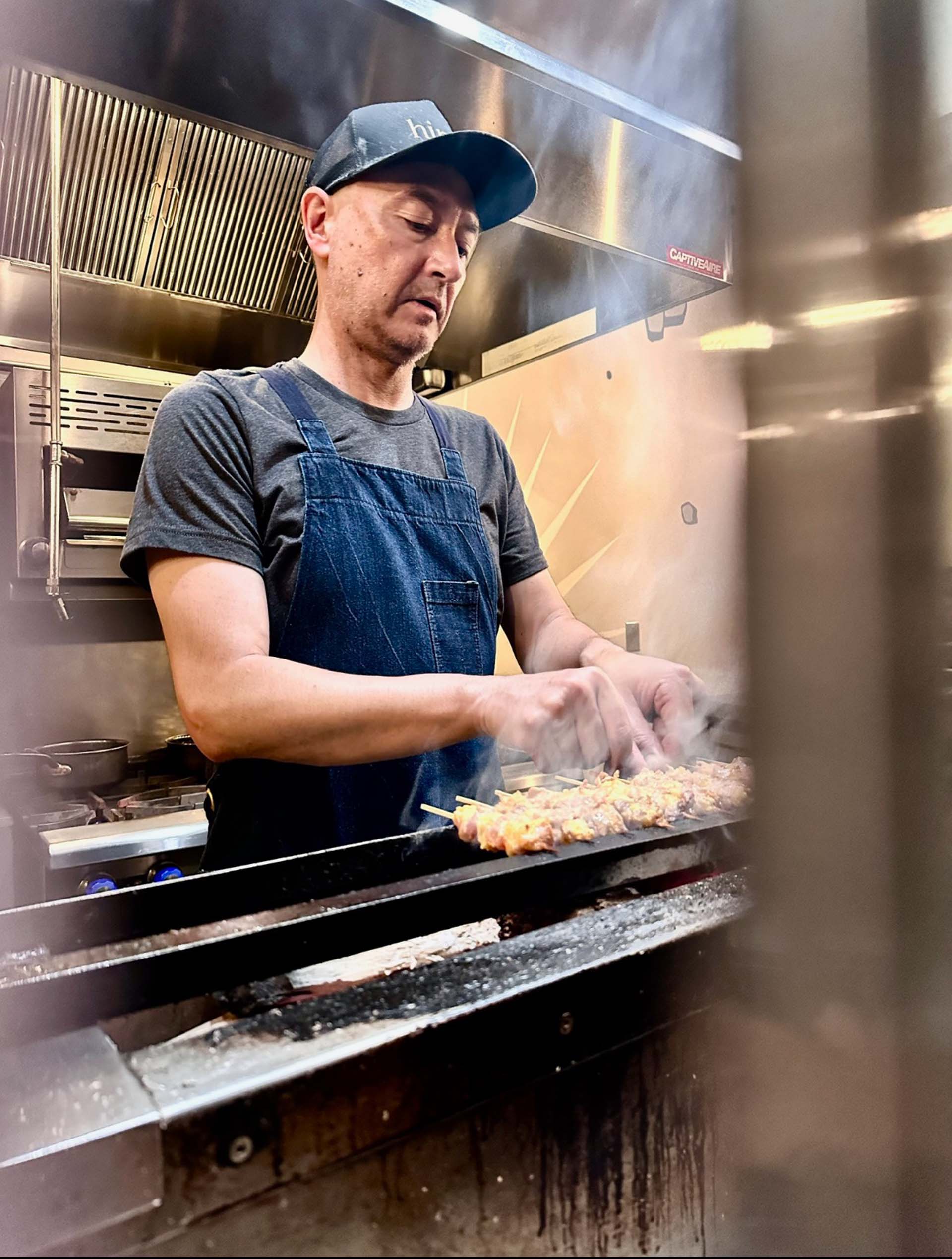 A yakitori chef in a baseball cap and apron turns chicken skewers over a smoky charcoal gril.