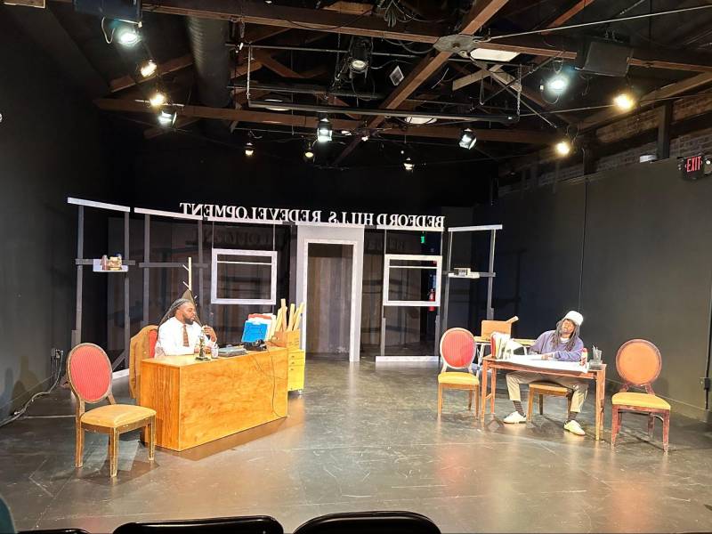 a stage with two desks and two Black men, actors, sitting behind them 