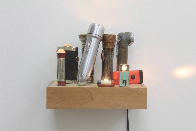 Nine flashlights of varying age turned on and arranged on a wooden shelf, extension cord extending from lower right corner