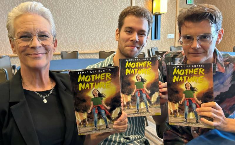 A white woman with cropped grey hair and silver rimmed glasses stands with two white men. All three hold up a comic book titled 'Mother Nature.' All are half-smiling.