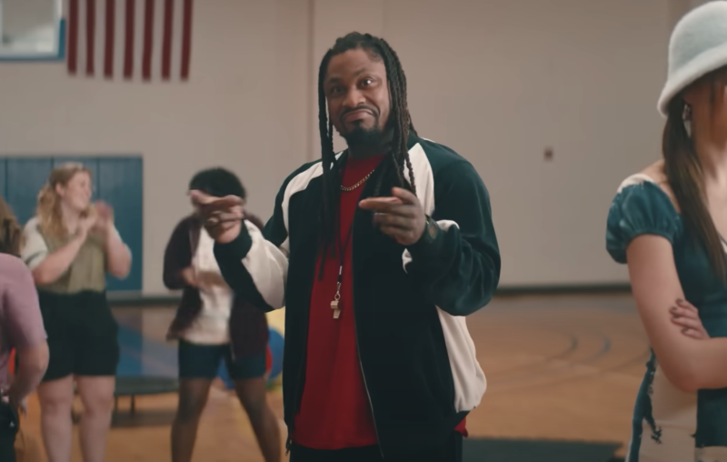 A Black man with long braids stands in the middle of a high school gym wearing sweats, smiling and gesturing. Students can be seen in the background.