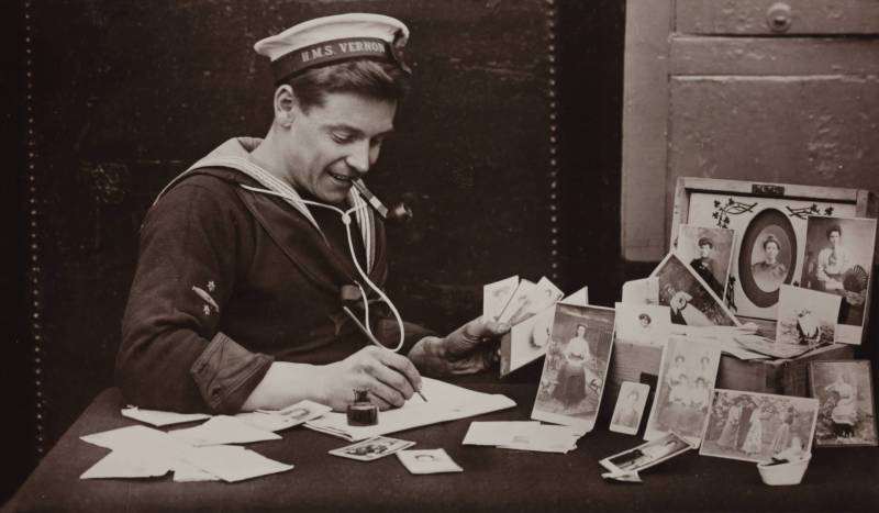 A 1940s-era sailor in uniform sits at a table writing letters, with a pipe in his mouth, surrounded by photographs of loved ones.