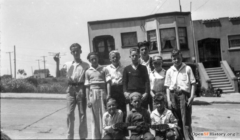 Ten small boys, between the ages of approximately 8 and 14 stand in the street in the 1920s. Most look sullen.