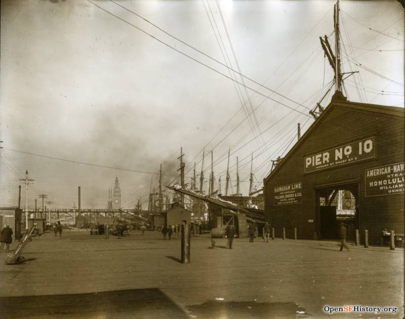 A black wooden shack marked PIER 10 sits before ships docked on the San Francisco Bay. The air is thick with smoke. The ferry building is visible off in the distance.
