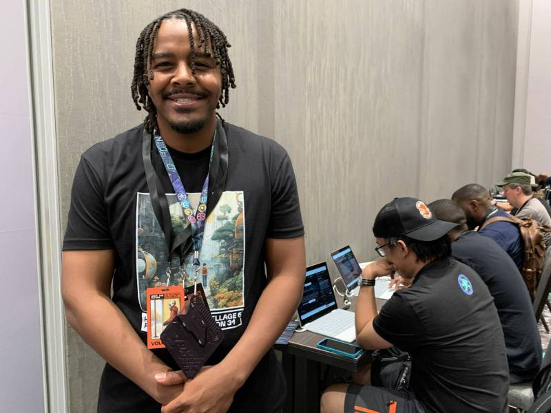 A smiling young Black man wearing a black t-shirt and two lanyards smiles warmly as a row of male computer users sit in a row behind him and stare at their laptops.