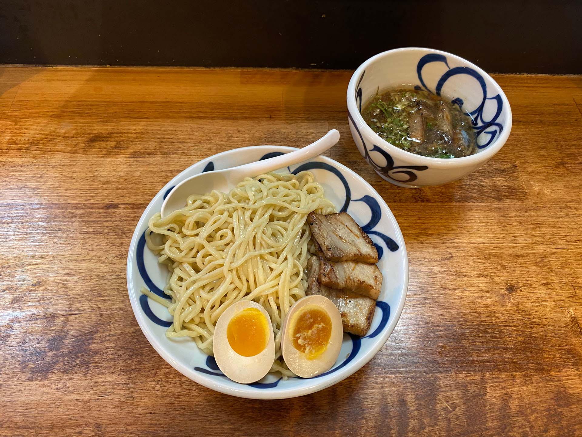 A bowl of cold ramen noodles topped with grilled pork belly and a halved soft-boiled egg, with dipping broth served in a separate small bowl on the side.