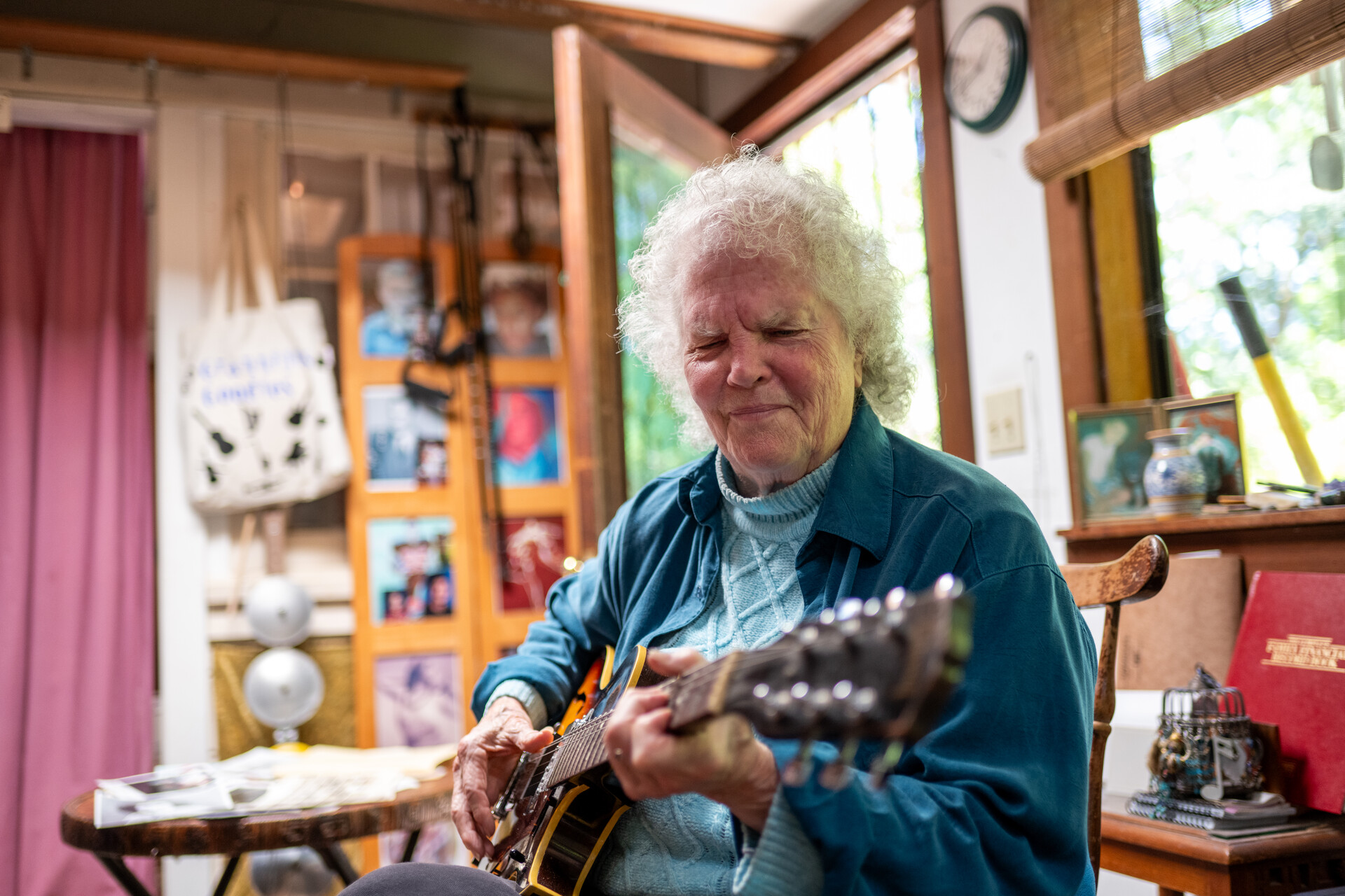 an older white woman with curly white hair smiles while sitting in her living room and playing guitar