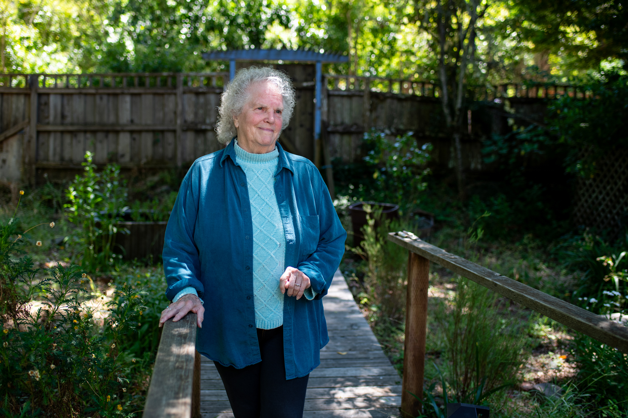 And older woman with short, curly gray hair smiles in leafy yard, one hand on wooden railing