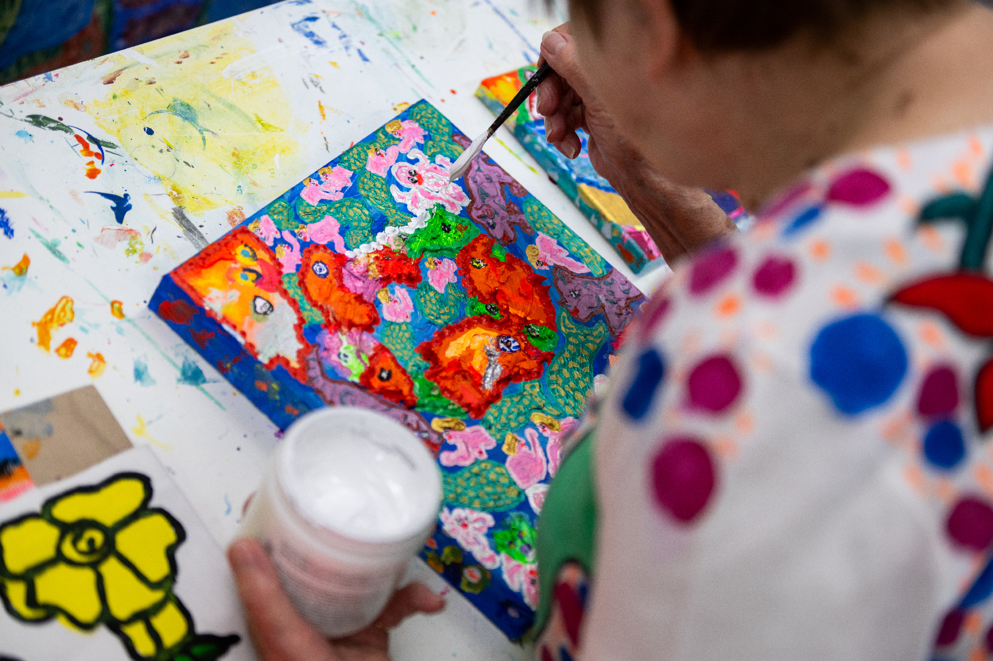 Over the shoulder shot of woman with white paint jar in left hand and paintbrush in right, dabbing thick dots onto a colorful small canvas