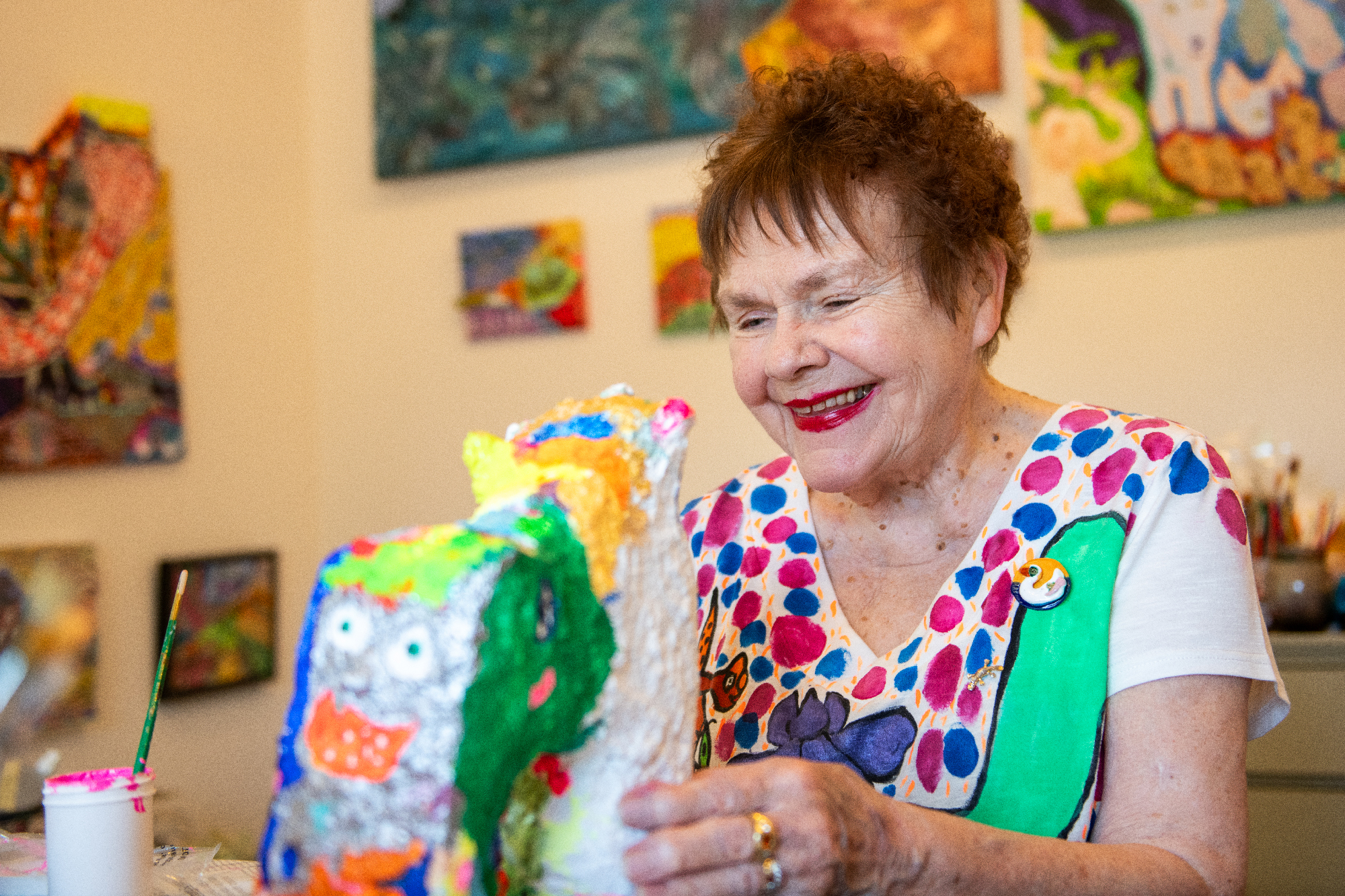 An older woman in a hand-painted shirt looks down at a colorful  sculpture with paintings on the wall behind her.