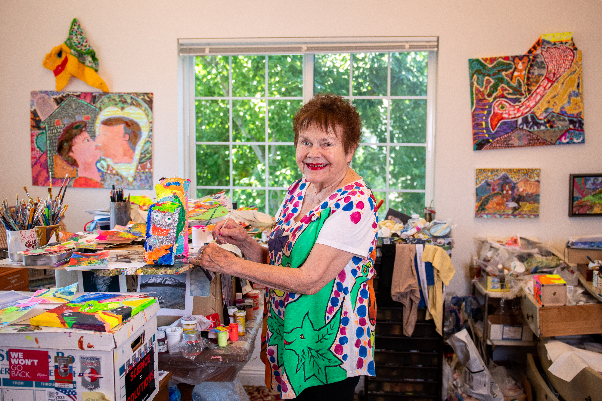 An older woman in a hand-painted shirt smiles while holding a brightly painted sculpture, surrounded by art supplies and artwork