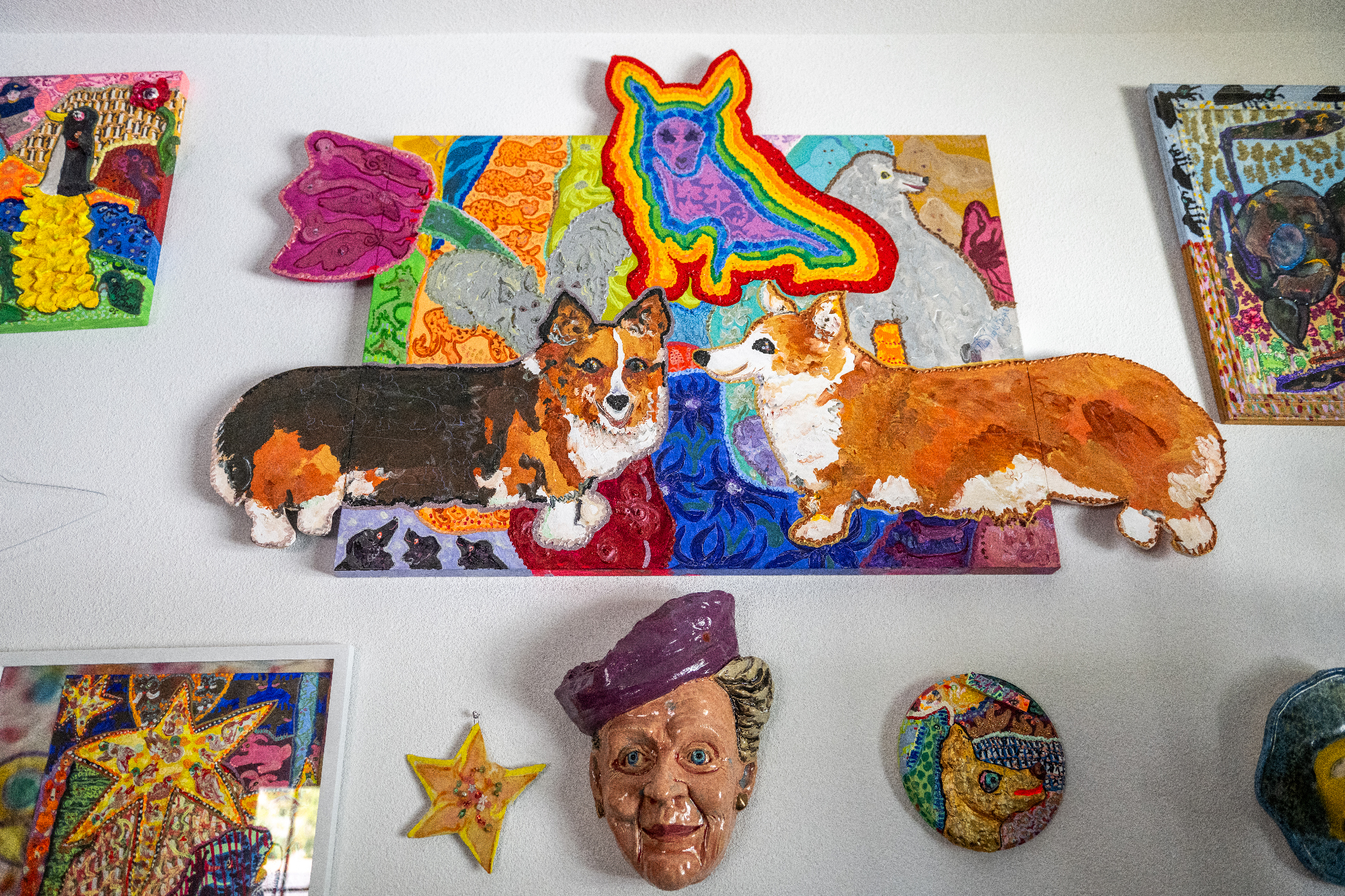 View of wall covered in colorful paintings and ceramic sculptures, including a painting of three Corgis with cut-out elements that extend dog bodies beyond rectangular canvas 