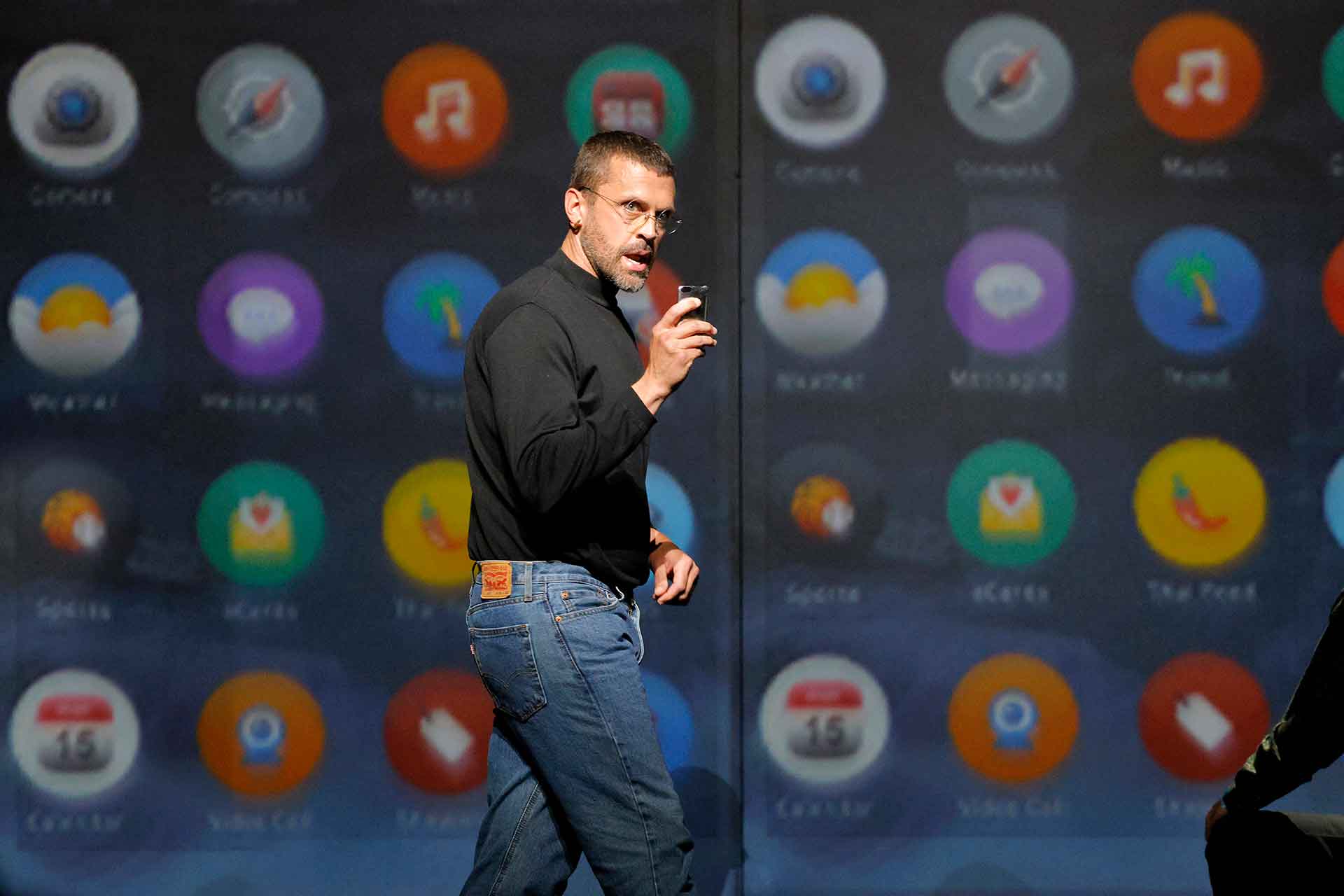 A man in a black turtleneck and jeans looks forward and to the right while holding a phone, against a backdrop of app icons