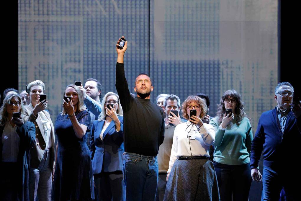 A man in a black turtleneck lifts a phone in the air as people dressed in 2007 corporate fashion stare at their own phones
