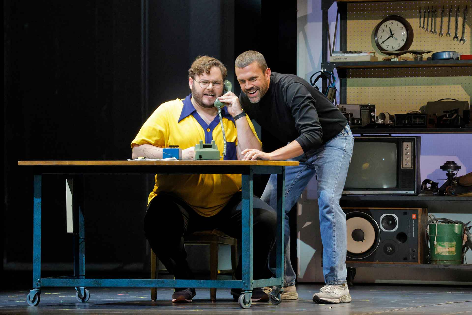 A man in a black turtleneck and jeans plays on a rotary phone with a man in scruffy hair, beard and yellow shirt at a workbench