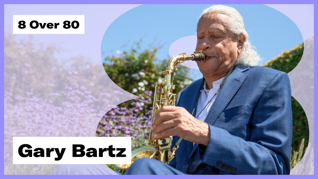 A man with white hair and a blue suit plays alto saxophone with foliage in the background.