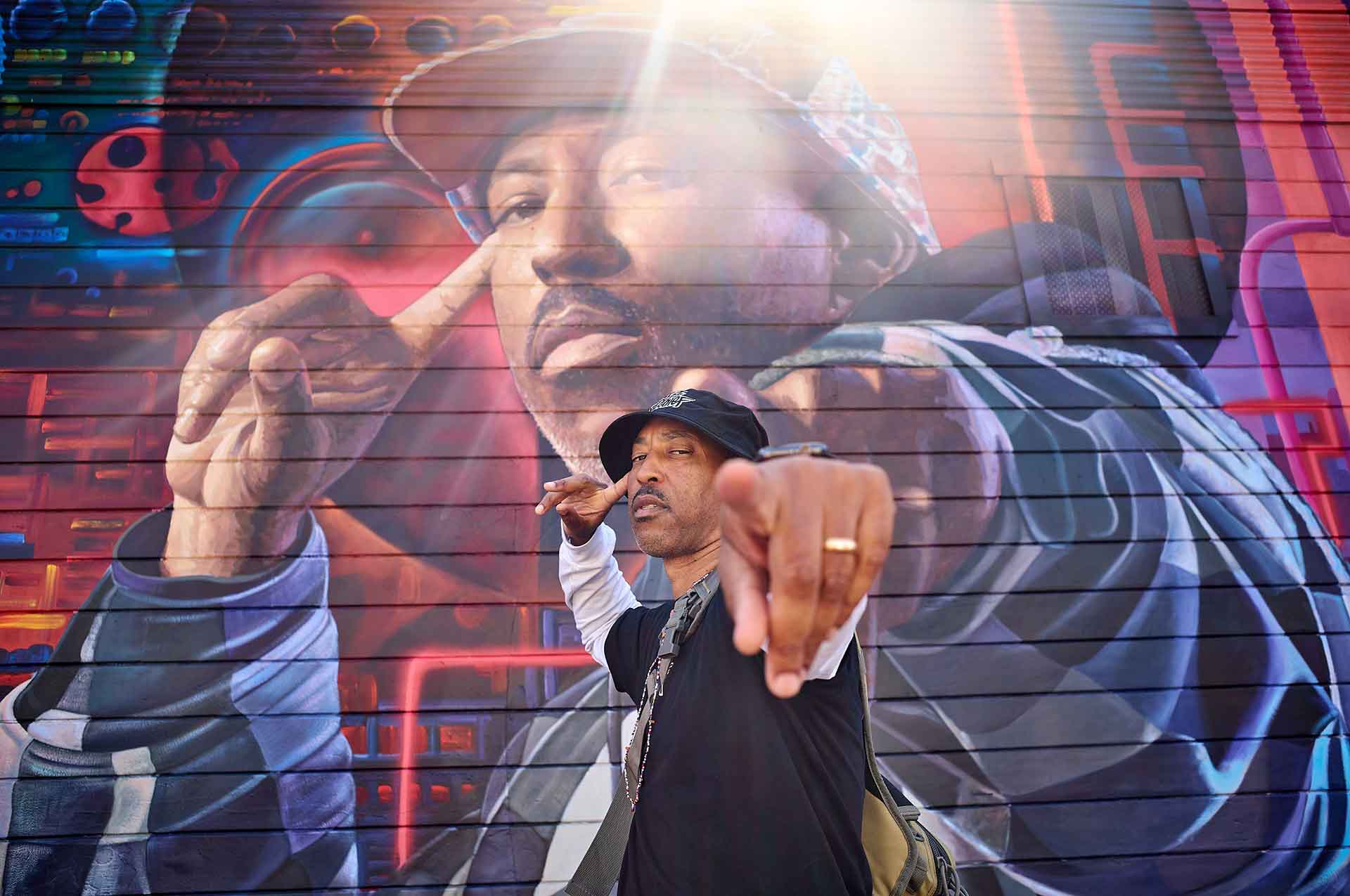 Phesto Dee of Souls of Mischief poses in front of his own likeness, painted as part of a mural in East Oakland.