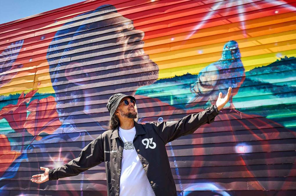 Opio of Souls of Mischief poses in front of his own likeness, painted as part of a mural in East Oakland.