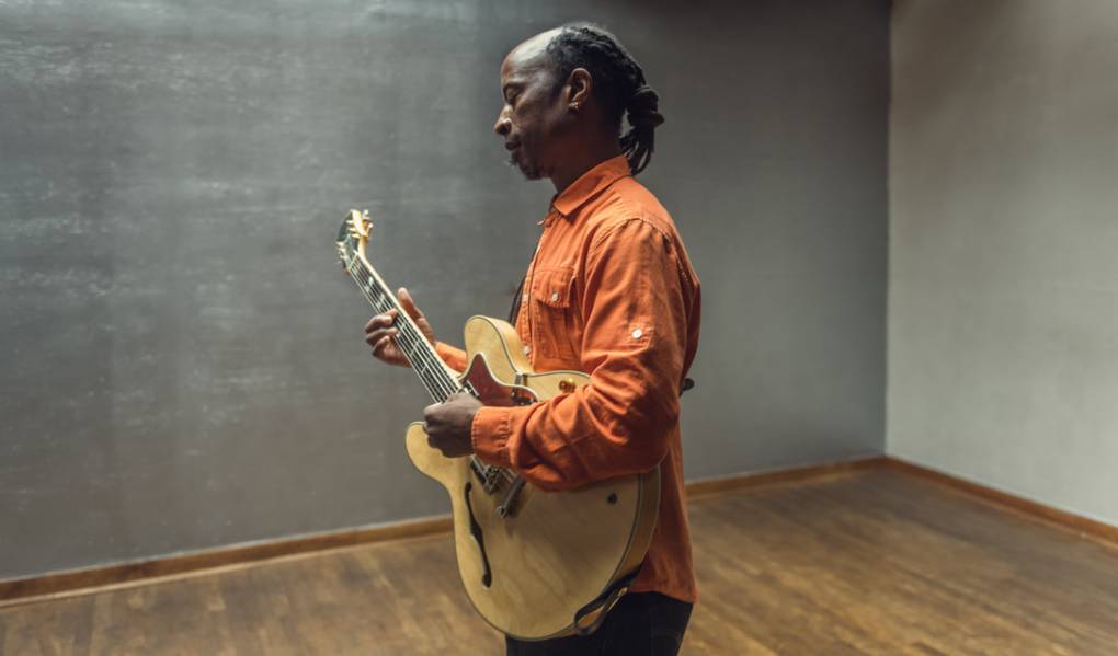a Black man wears a button down orange shirt and holds a guitar standing against a gray wall