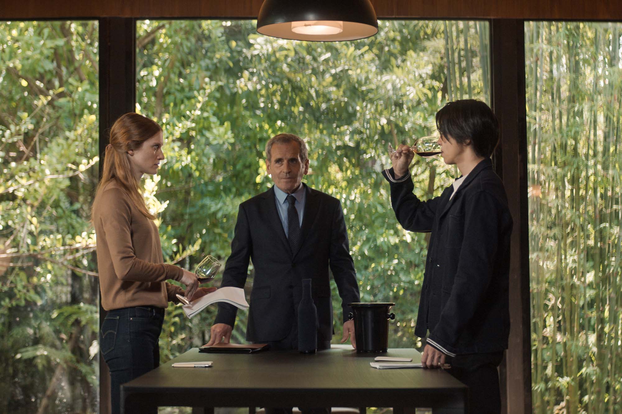A white woman and Japanese woman taste wine while standing across a table from each other. A man in a suit looks on; the backdrop outside the window is very verdant and green.