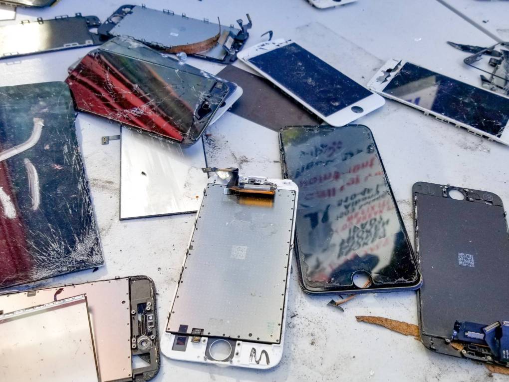 Close-up of a large number of broken cellphones, including cracked cellphone screens with exposed wiring, on a white surface.