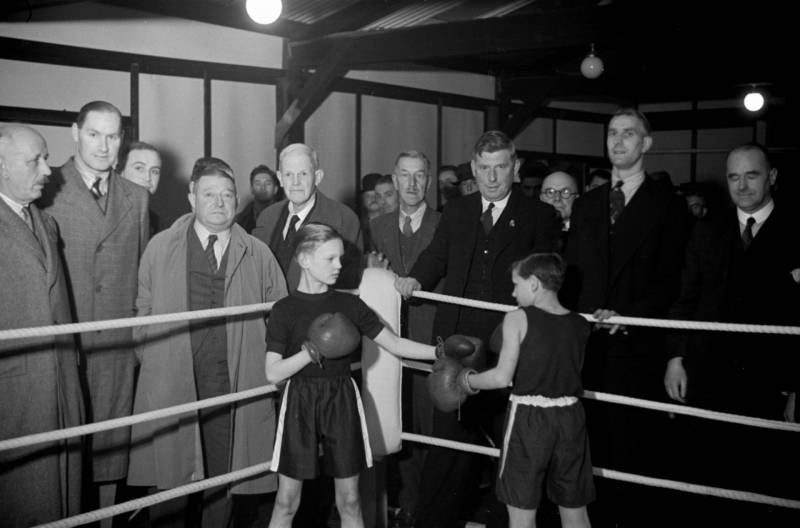 Two small boys boxing inside a ring surrounded by middle aged men wearing overcoats and suits.