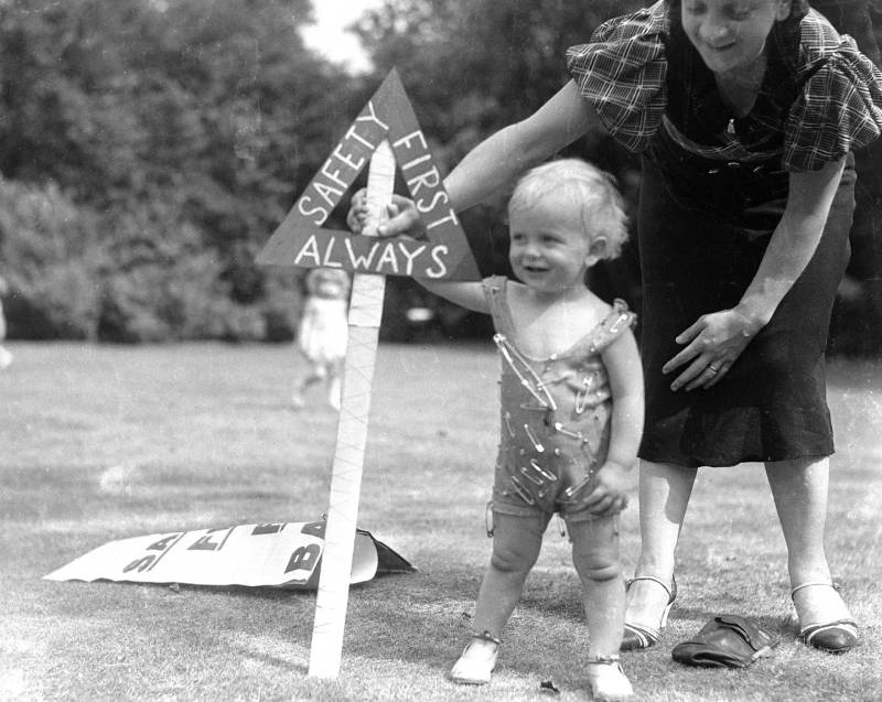 A toddler wearing a onesie covered in safety pins stands outside next to a sign that reads Safety First. A woman stands behind the child.