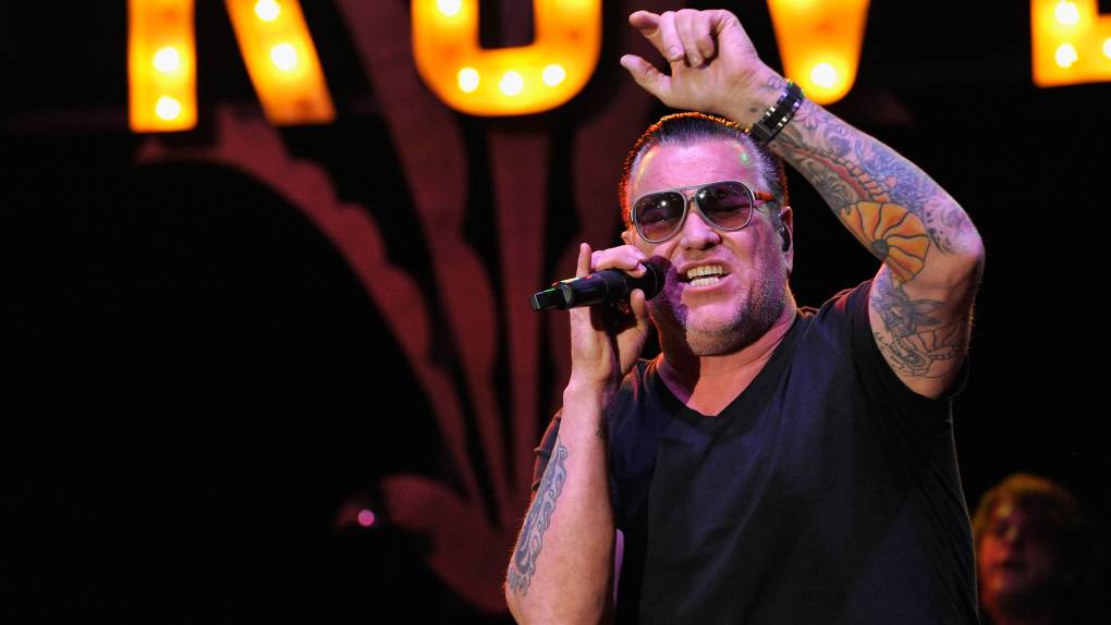 a man in sunglasses, black t-shirt and tattoos sings into a microphone